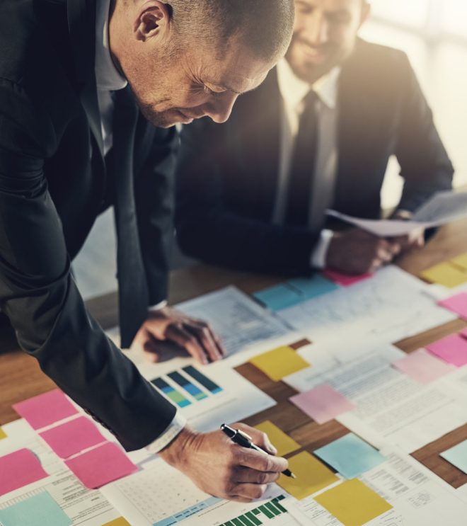Business men, partner and writing notes with documents, graphs and financial planning for architecture. People, happy and paperwork with blueprint, charts and sign for property expansion at company.